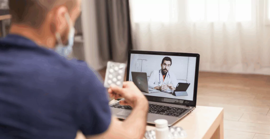 Homem segurando uma cartela de medicamente de frente para um notebook que exibe na tela um médico